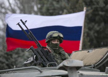 A pro-Russian man (not seen) holds a Russian flag behind an armed servicemen on top of a Russian army vehicle outside a Ukrainian border guard post in the Crimean town of Balaclava March 1, 2014. Ukraine accused Russia on Saturday of sending thousands of extra troops to Crimea and placed its military in the area on high alert as the Black Sea peninsula appeared to slip beyond Kiev's control. REUTERS/Baz Ratner (UKRAINE - Tags: MILITARY POLITICS CIVIL UNREST)