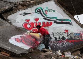 TOPSHOT - A man draws a slogan in Arabic reading "we still want freedom" as part of a graffiti mural commemorating the ninth anniversary of uprising that led to the Syrian conflict (March 15), showing a dove holding an olive branch in its beak flying over a Syrian opposition flag in the shape of the eastern Arabic numeral "9" while being targeted by the silhouette of a military aircraft with the Arabic word "years" below as silhouettes of children stand by a border fence and a tent with the letters "UN", drawn on the collapsed roof of a heavily damaged building in the town of Binnish in the northwestern Idlib province on March 13, 2020. (Photo by Omar HAJ KADOUR / AFP) (Photo by OMAR HAJ KADOUR/AFP via Getty Images)